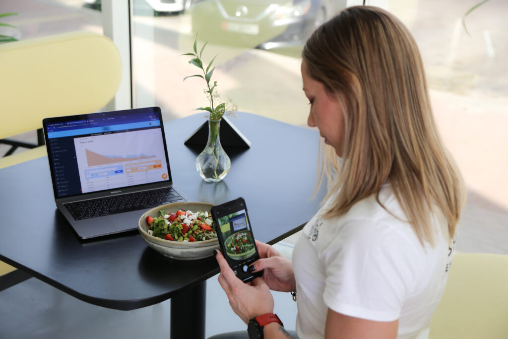 female influencer taking a picture of her salad bowl with mobile phone. laptop on the table displaying results graph